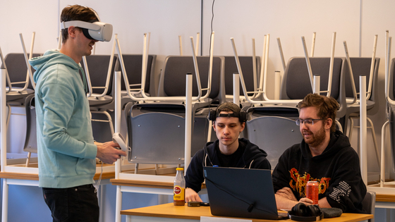 Mann står med VR-headset, mens to personer sitter ved siden av ved et bord