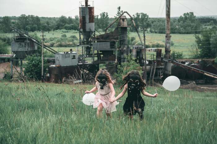 Two young girls on a field with ballons, gas masks covering their faces.