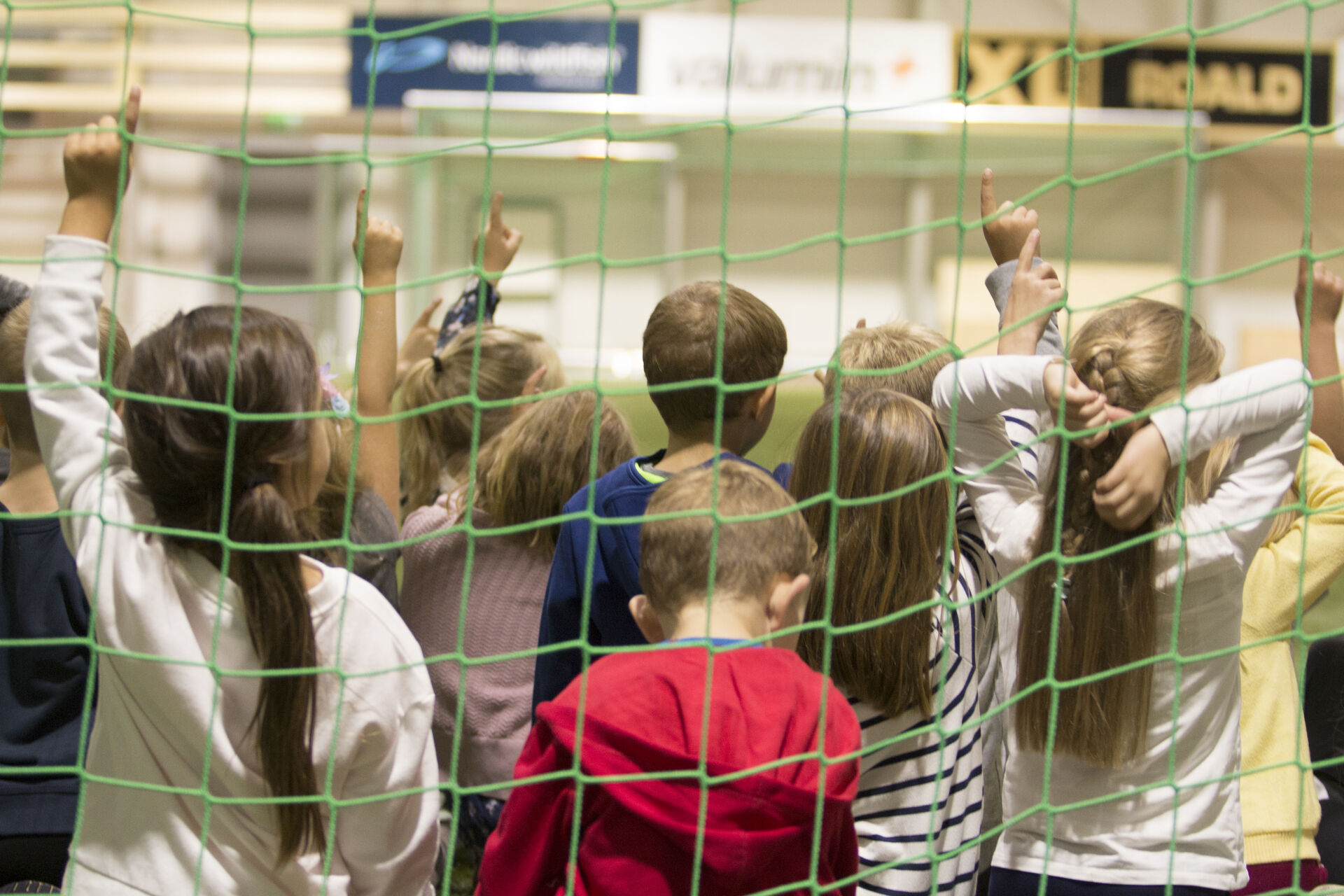 The picture shows the back of the heads of a group of children placed in front of a net in a sports hall